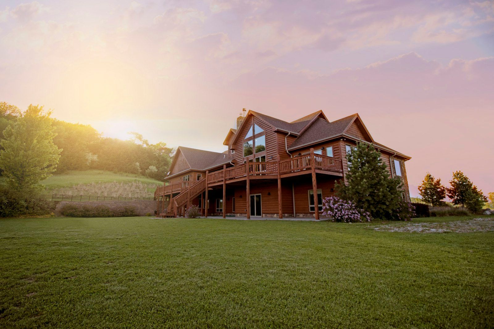 A wood-sided two-story home with a wraparound deck and a wide green lawn at sunset